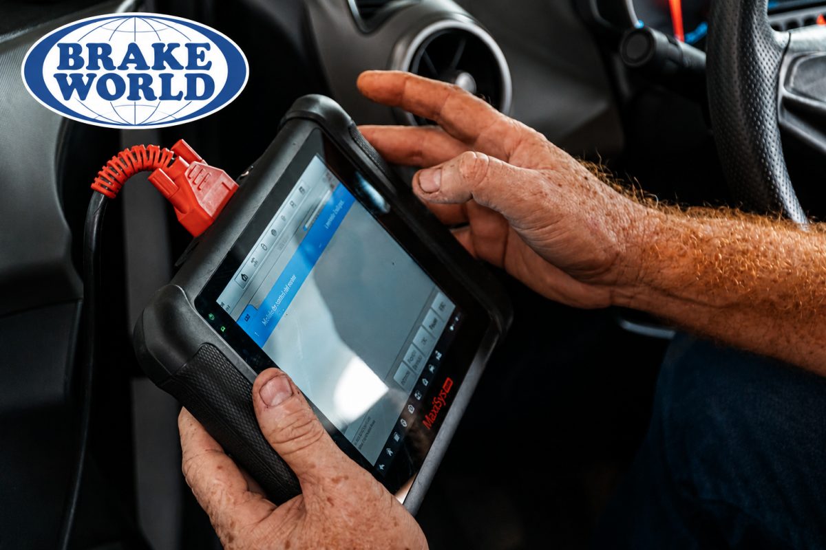 A mechanic's hands hold an automotive diagnostic tablet connected by a red cable inside a vehicle.